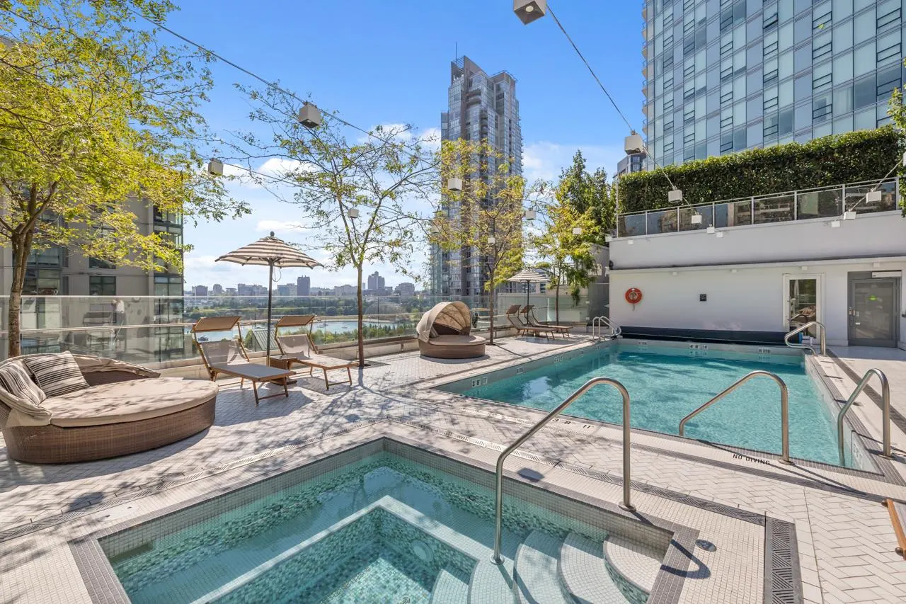 Rooftop pool area with a hot tub, lounge chairs, and a view of the city skyline on a sunny day.