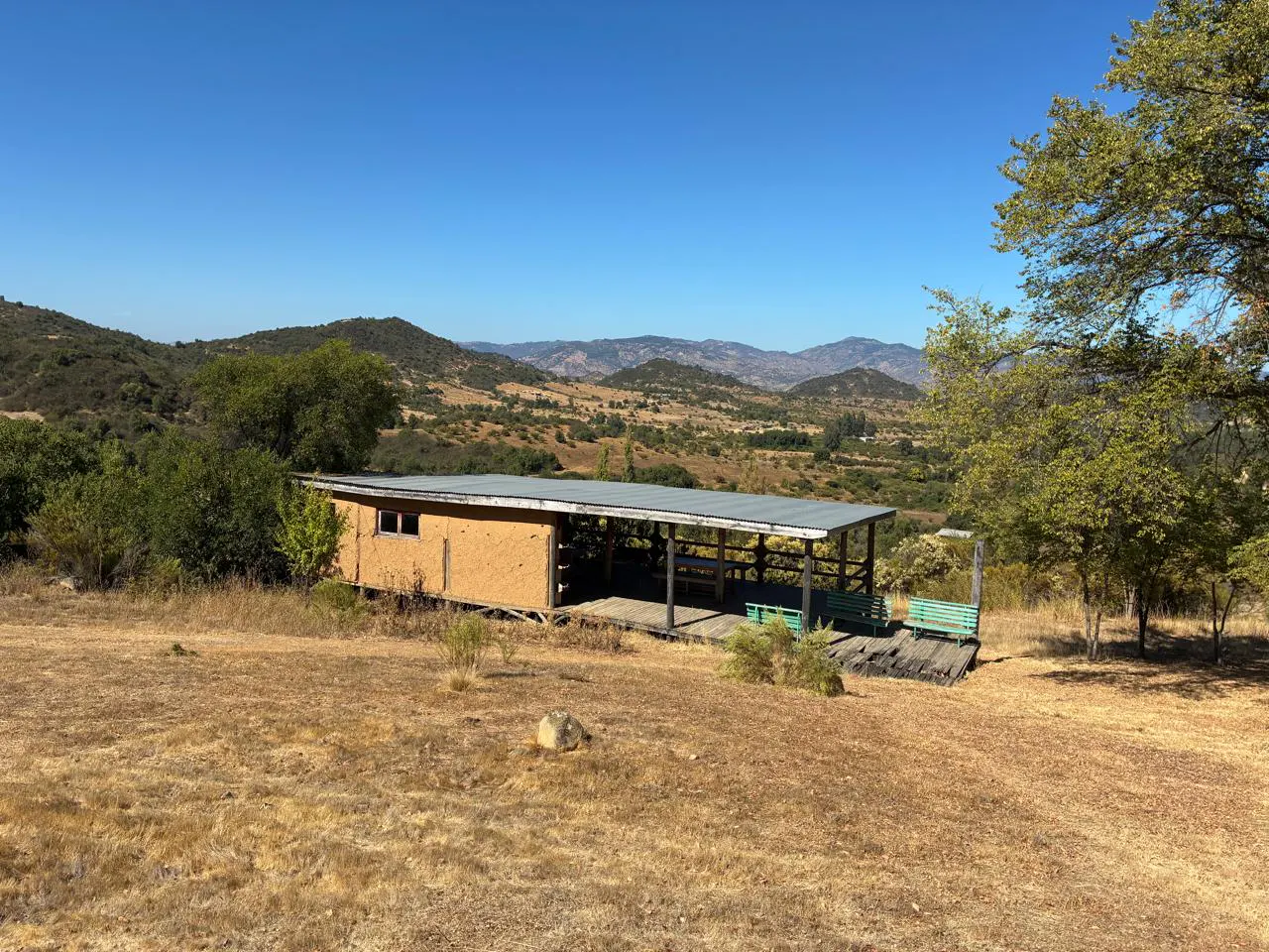 A small, tan house with a covered porch sits in a dry, grassy field with mountains in the background under a clear blue sky.