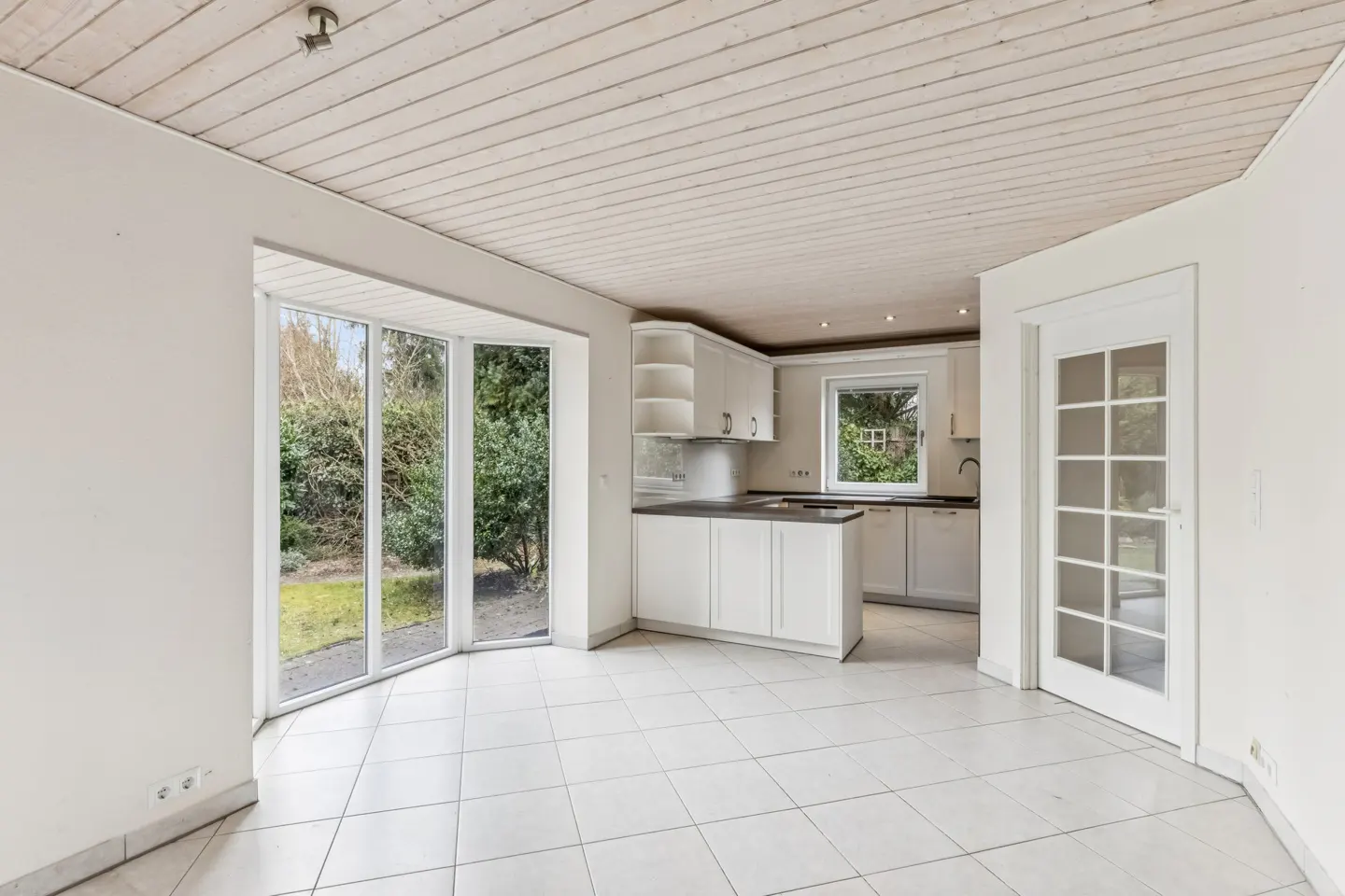 Bright, empty kitchen with white cabinets, tile floor, and a bay window overlooking a green garden. A white door with glass panes is on the right.