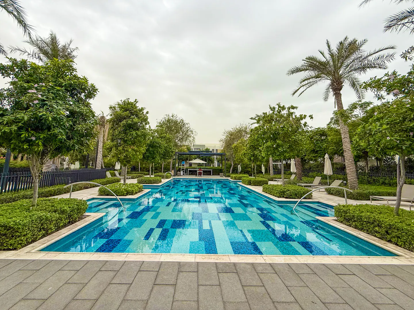 A rectangular swimming pool with blue tiles, surrounded by trees and lounge chairs, under a cloudy sky.