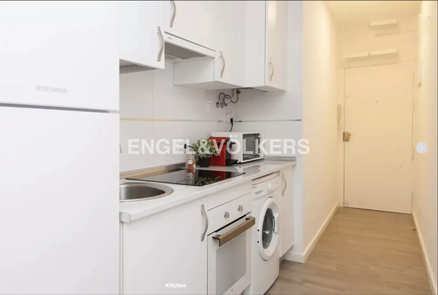 A bright, all-white kitchen with appliances, cabinets, and a hallway leading to a white door.