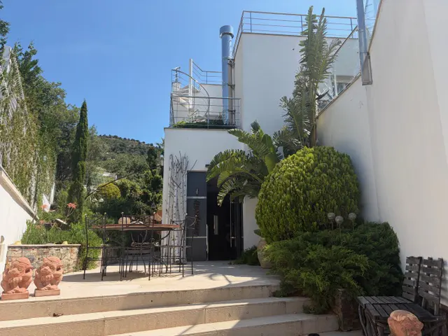 Exterior view of a modern white house with a rooftop terrace and spiral staircase. A patio with a table and chairs is surrounded by lush greenery.
