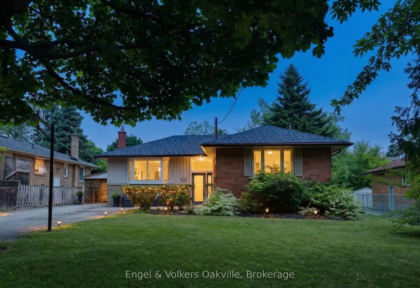 Exterior view of a brick and siding house with a dark roof, green lawn, and trees at dusk. Lights illuminate the front yard.