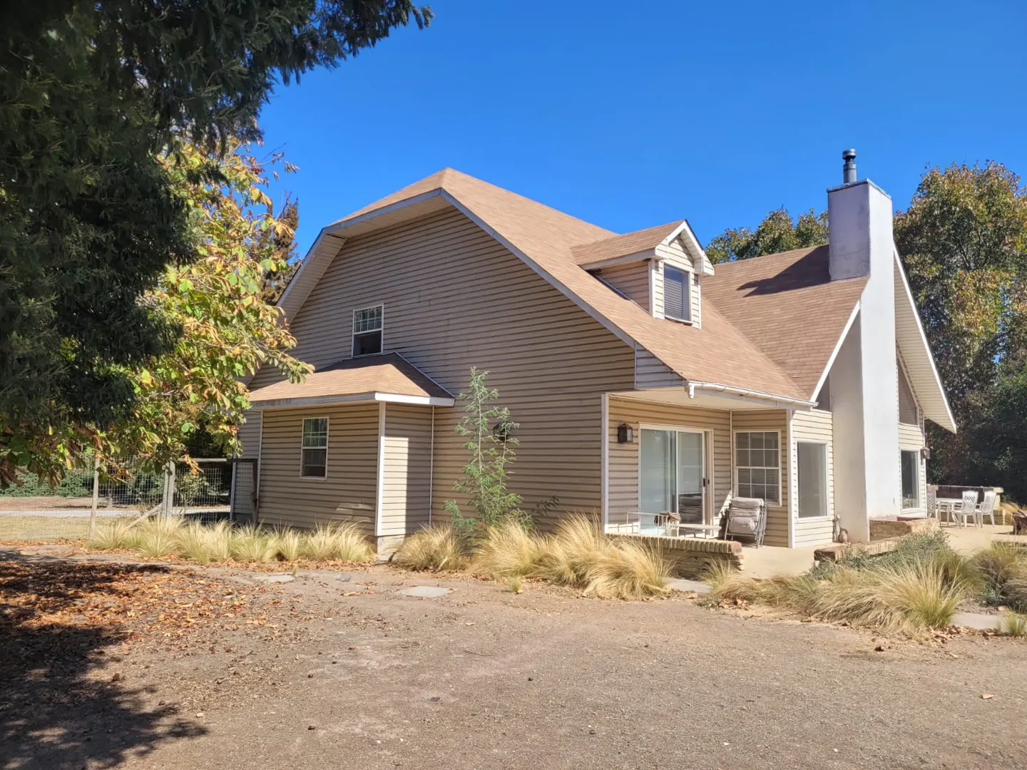 Tan two-story house with a brown roof and white chimney under a blue sky. Trees surround the house.