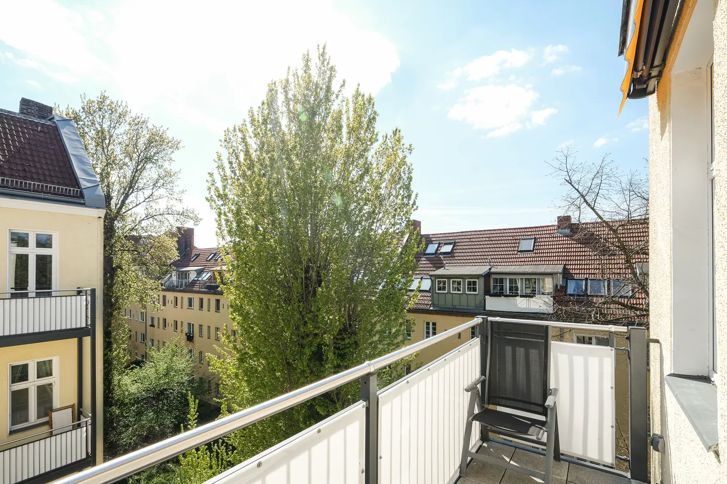 View from a balcony with a metal railing, a chair, and a view of trees and buildings on a sunny day.