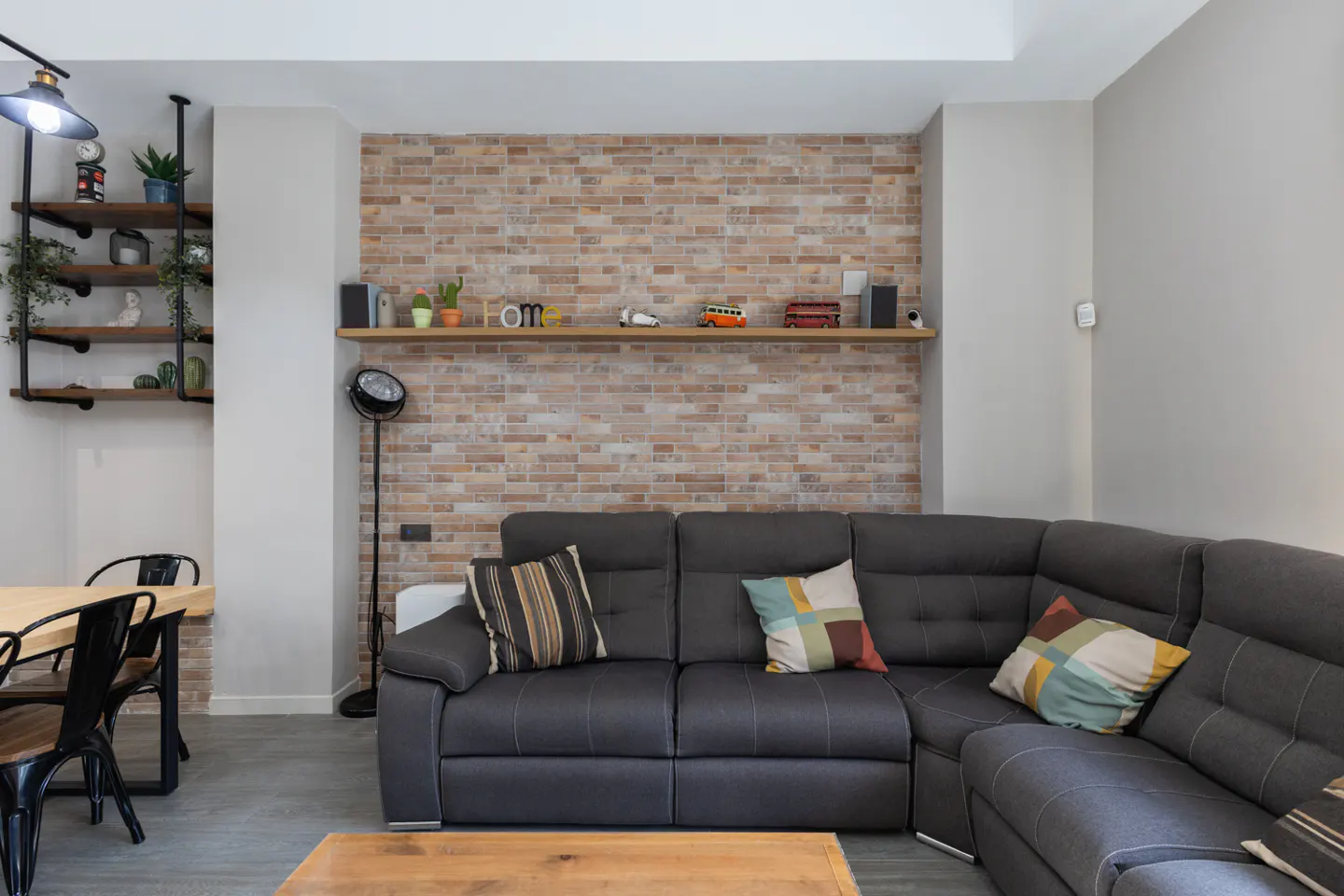 Living room with a gray sectional sofa, brick wall, and wooden shelves with plants and decor. A wooden table and black chairs are visible.