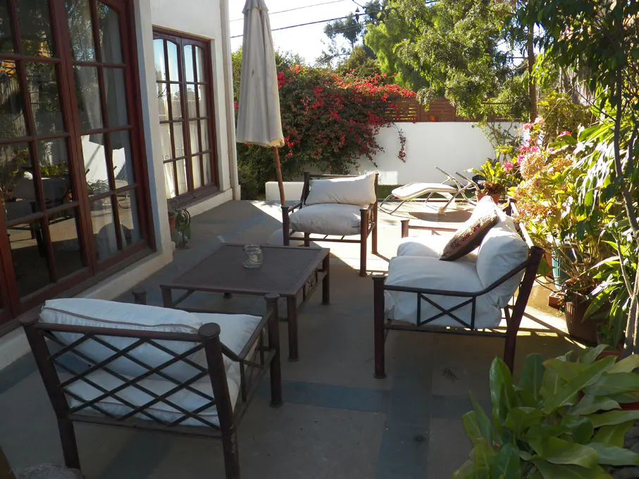 Outdoor patio with brown metal furniture, white cushions, and a closed umbrella. Red flowers and green trees in the background.
