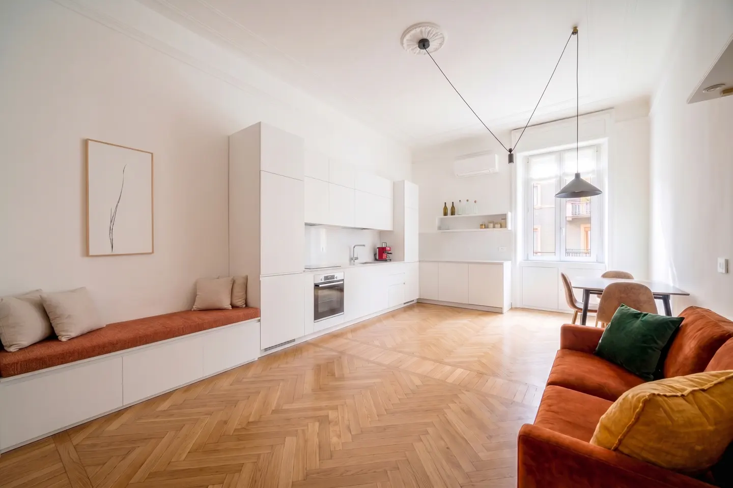 Modern kitchen and living area with white cabinets, herringbone wood floors, a rust-colored sofa, and built-in bench seating.