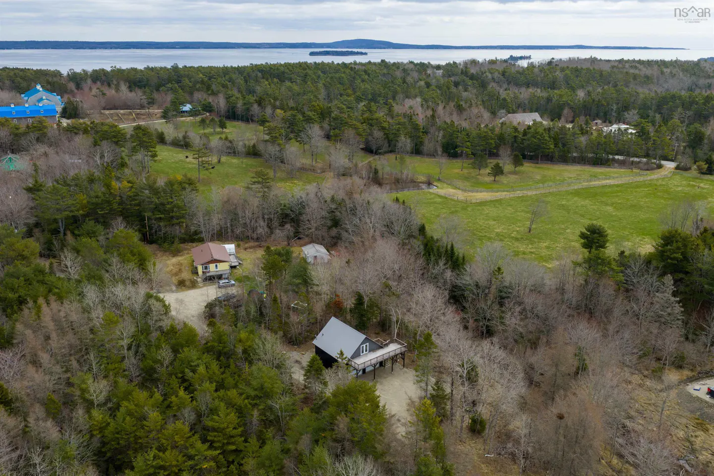 Aerial view of a wooded property with a gray house, green fields, and a lake in the background.