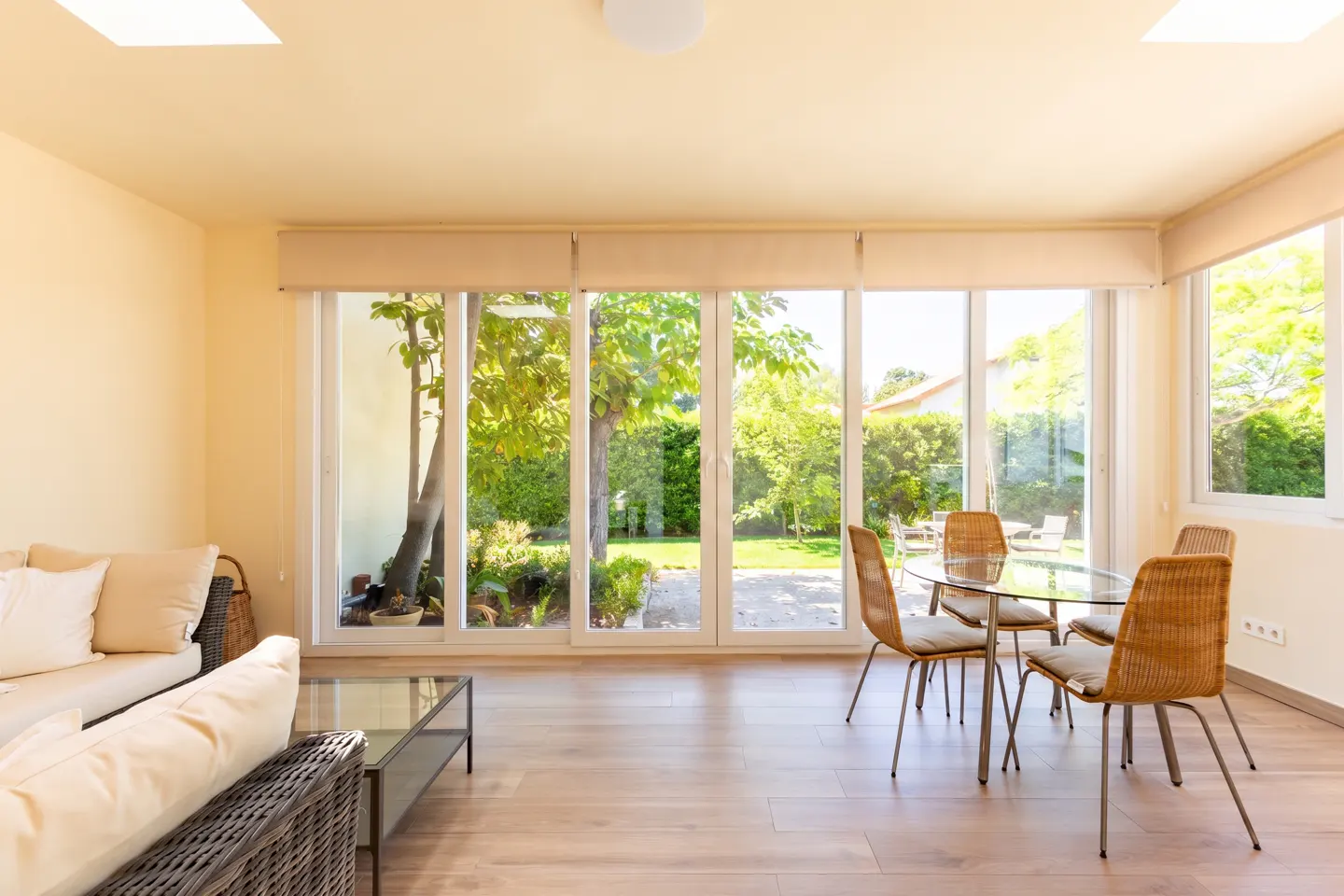 Bright living room with a glass table, wicker chairs, and a sofa. Large windows overlook a green garden.