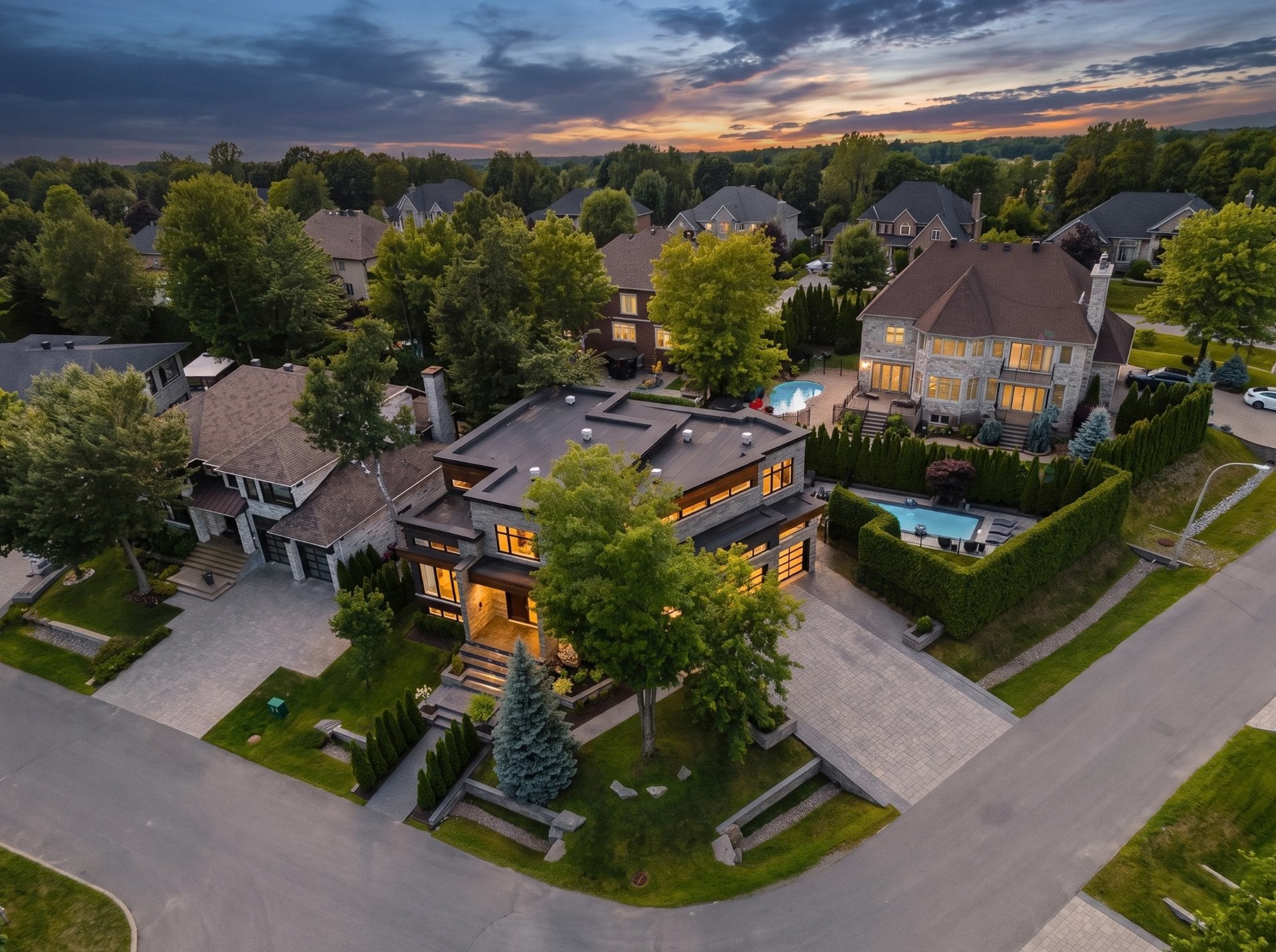 Aerial view of a modern luxury home with a flat roof and glowing interior lights at sunset in a suburban neighborhood.