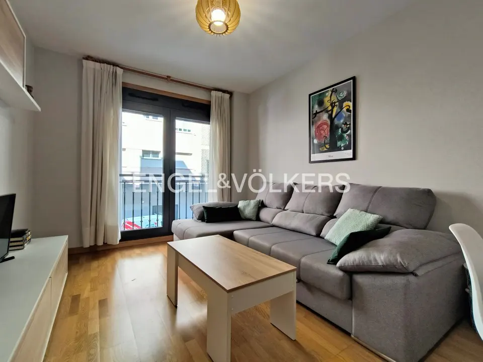 Living room with hardwood floors, a gray sectional sofa, and a light wood coffee table. Balcony doors with white curtains are in the background.