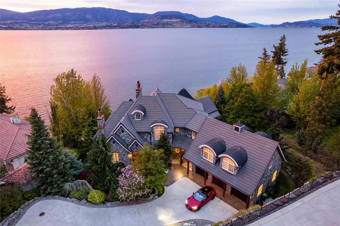 Aerial view of a large stone house with a gray roof, surrounded by trees, overlooking a lake and mountains at sunset. A red car is parked in the driveway.