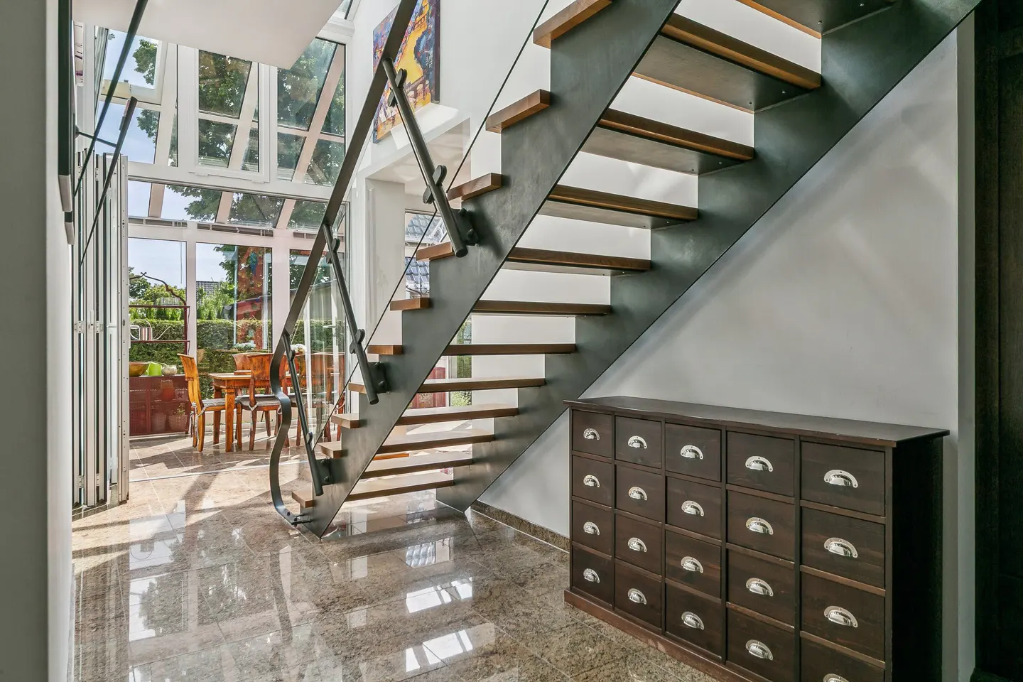 Modern staircase with wooden treads and metal frame, next to a dark wood apothecary cabinet on a polished stone floor.