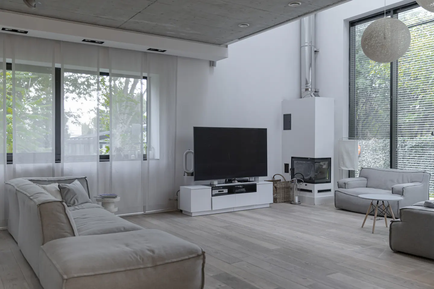 Bright, modern living room with a gray sectional sofa, large TV, and white fireplace. Large windows with sheer curtains let in natural light.