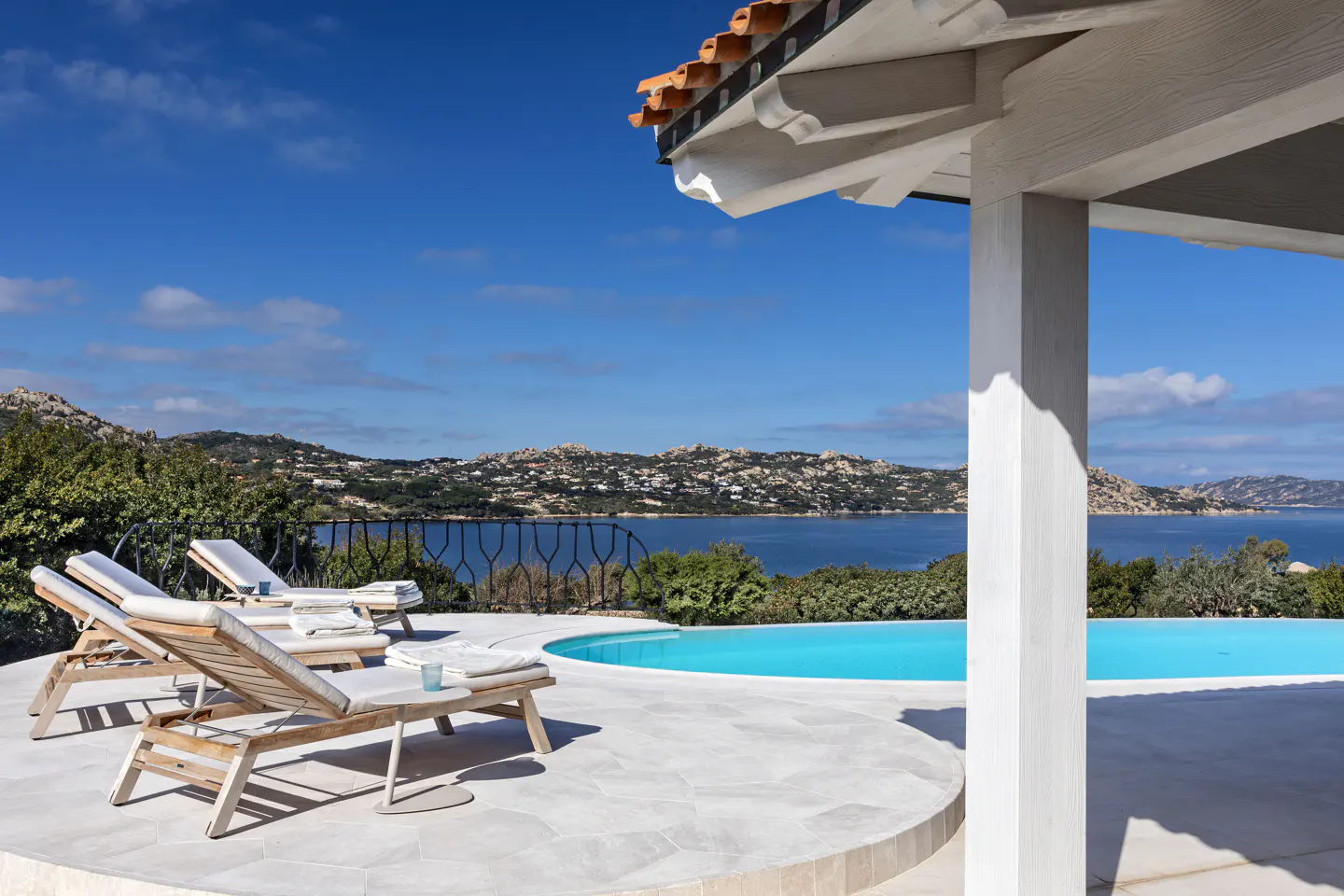 Luxury villa with pool overlooking the ocean. Lounge chairs sit on a white patio under a roof with orange tiles.