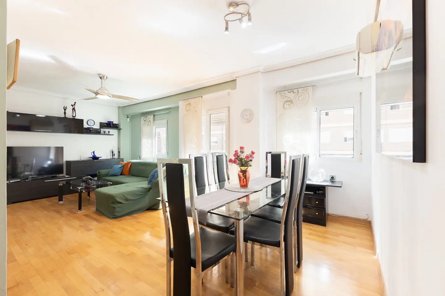 Bright living and dining room with hardwood floors, a green sectional sofa, and a glass dining table with black chairs.