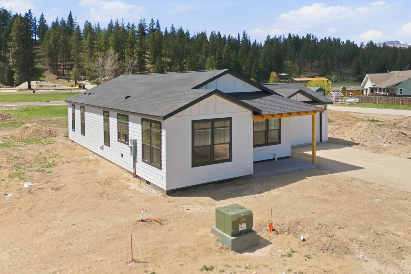 A modern, white house with black trim and a gray roof sits on a dirt lot with trees in the background.