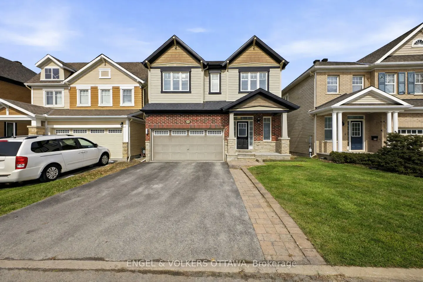 Two-story house with beige siding, brick accents, and a gray garage door. A long driveway leads to the house, with green lawns on either side.