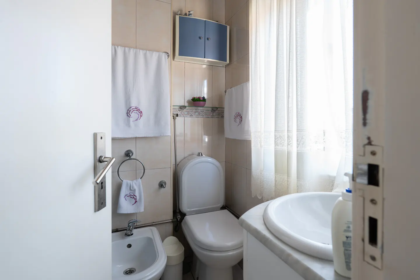 A bright bathroom with a toilet, bidet, sink, and white towels with a purple design. A blue cabinet hangs above the toilet. The door is ajar.