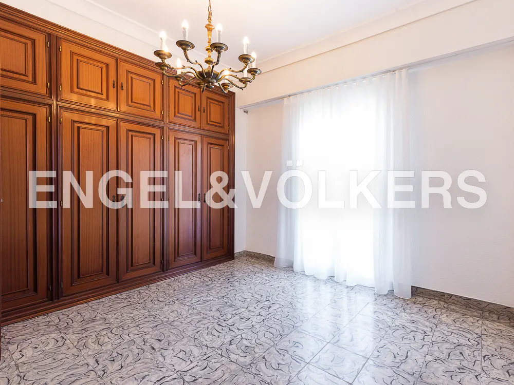 A bedroom with a large wooden wardrobe, a chandelier, and patterned tile flooring. A window with white curtains is visible.