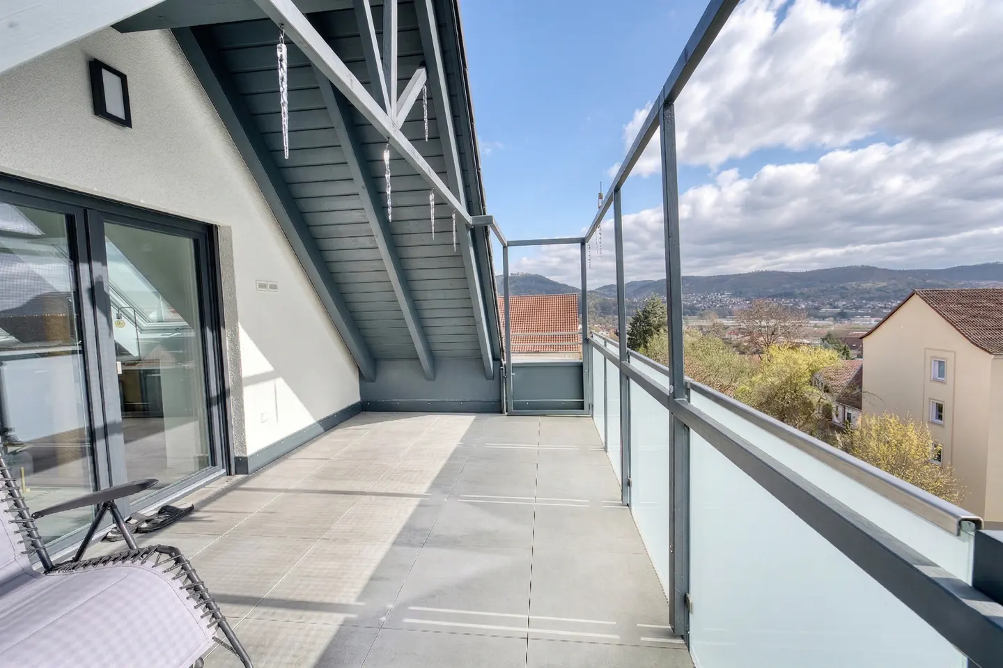Balcony with gray tile floor, glass railing, and gray roof. A chair sits near a sliding glass door, with a view of houses and hills in the background.
