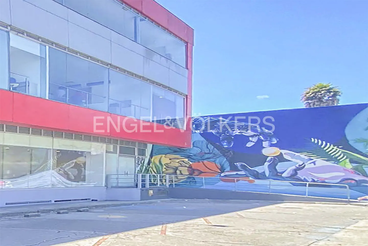 Exterior of a modern building with red accents and large windows, next to a colorful mural under a blue sky.