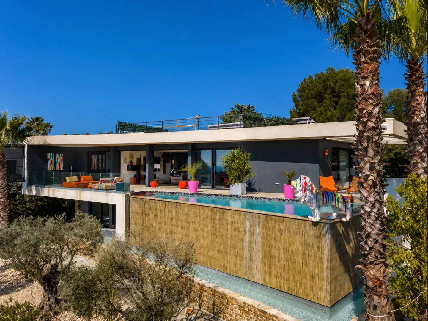 Modern villa with an infinity pool, a colorful lion sculpture, and a terrace under a clear blue sky.