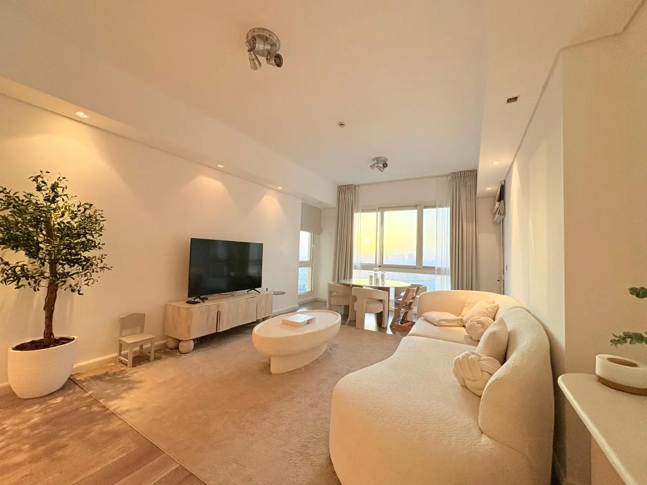 A bright, modern living room with a large, curved white sofa, oval coffee table, and a TV on a light wood cabinet. A potted tree and a small chair add a touch of nature.