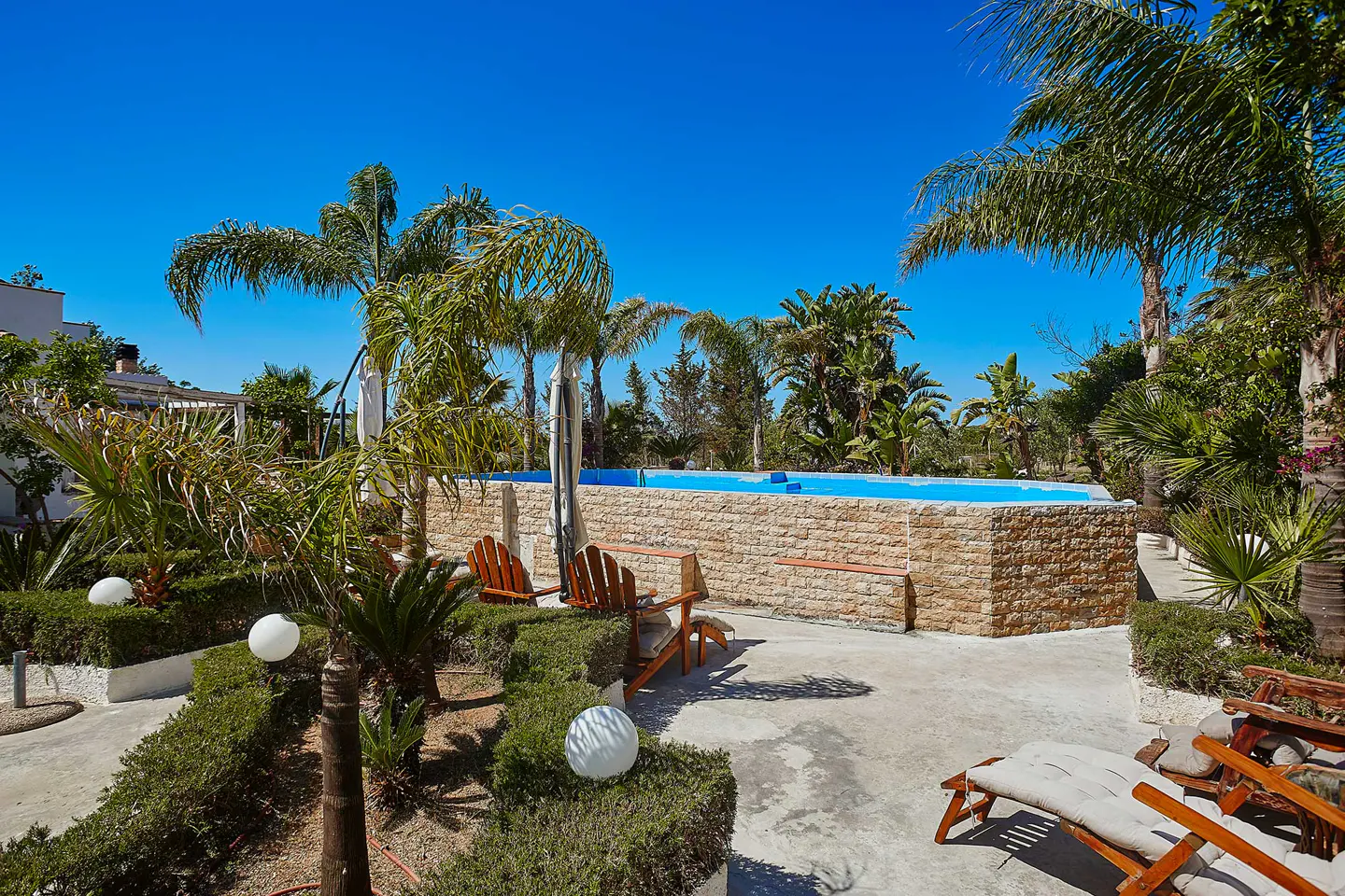 Raised swimming pool with stone walls, surrounded by palm trees, manicured hedges, and lounge chairs under a clear blue sky.