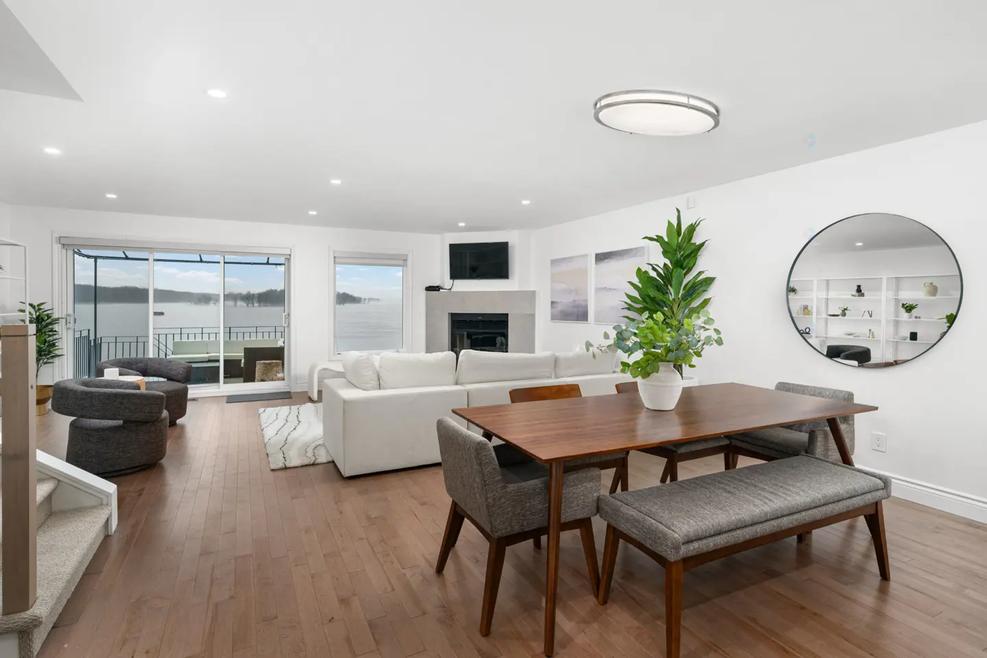 Bright, open-concept living and dining area with hardwood floors. White walls, sofa, and fireplace. Lake view through sliding glass doors.