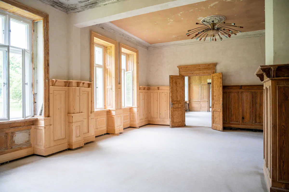Empty room with light wood paneling, large windows, and an open doorway. A unique chandelier hangs from the ceiling.