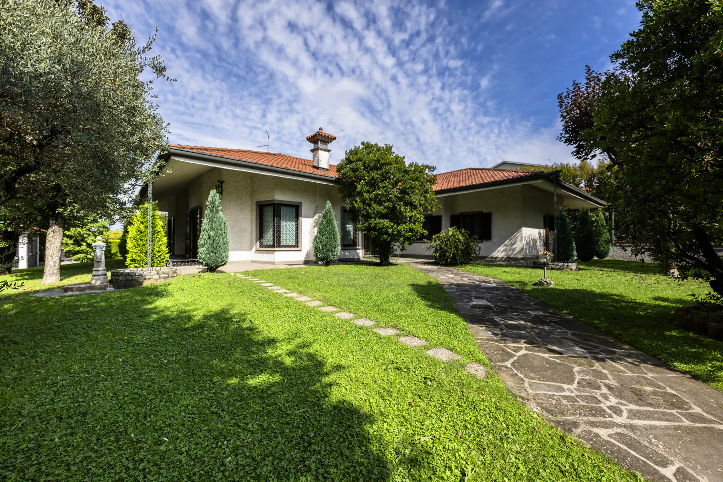 A single-story house with a red tile roof, beige walls, and a stone walkway leading to the entrance. Green lawn and trees surround the house.