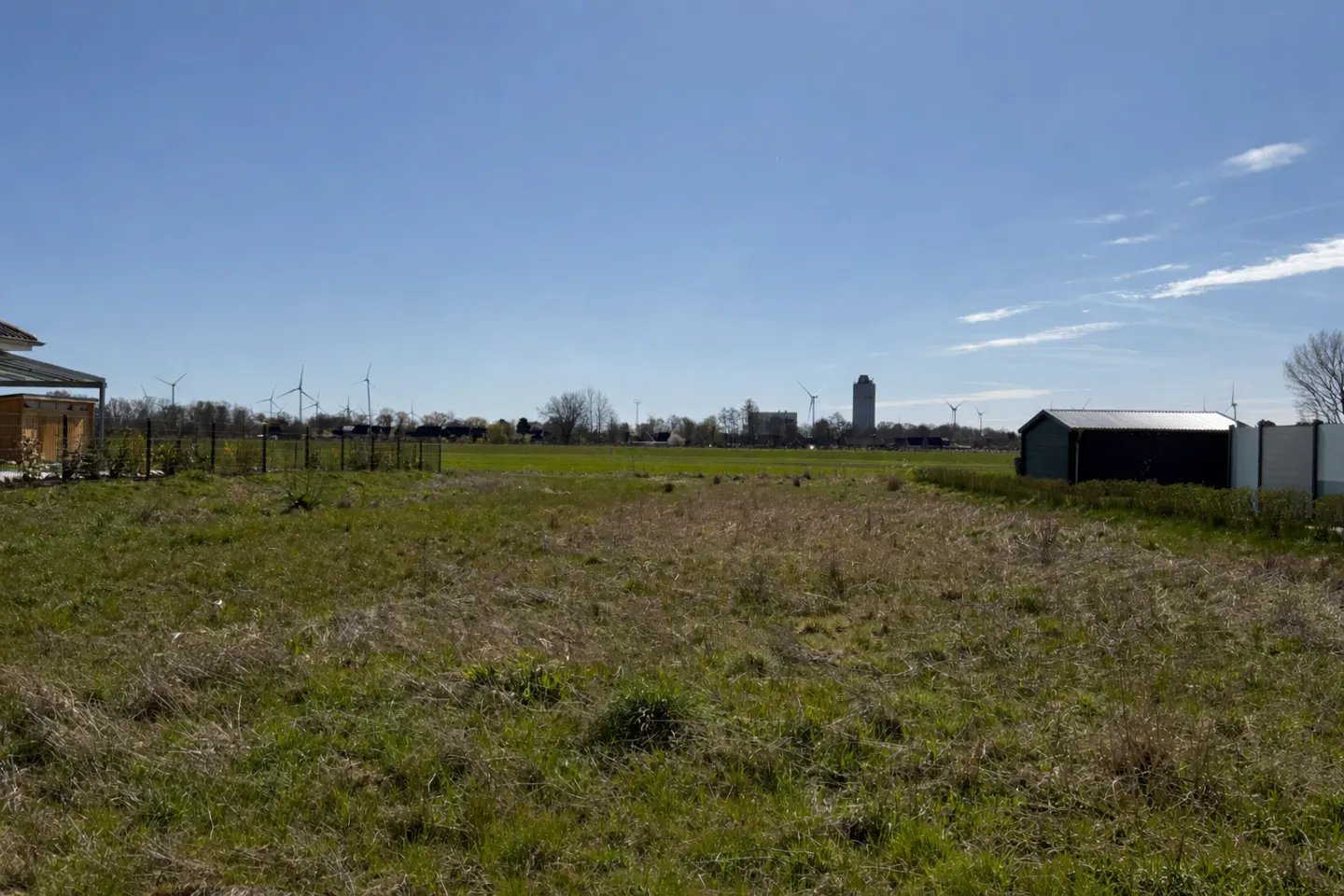 A grassy field under a blue sky, with wind turbines and a building in the distance. A shed is on the right.