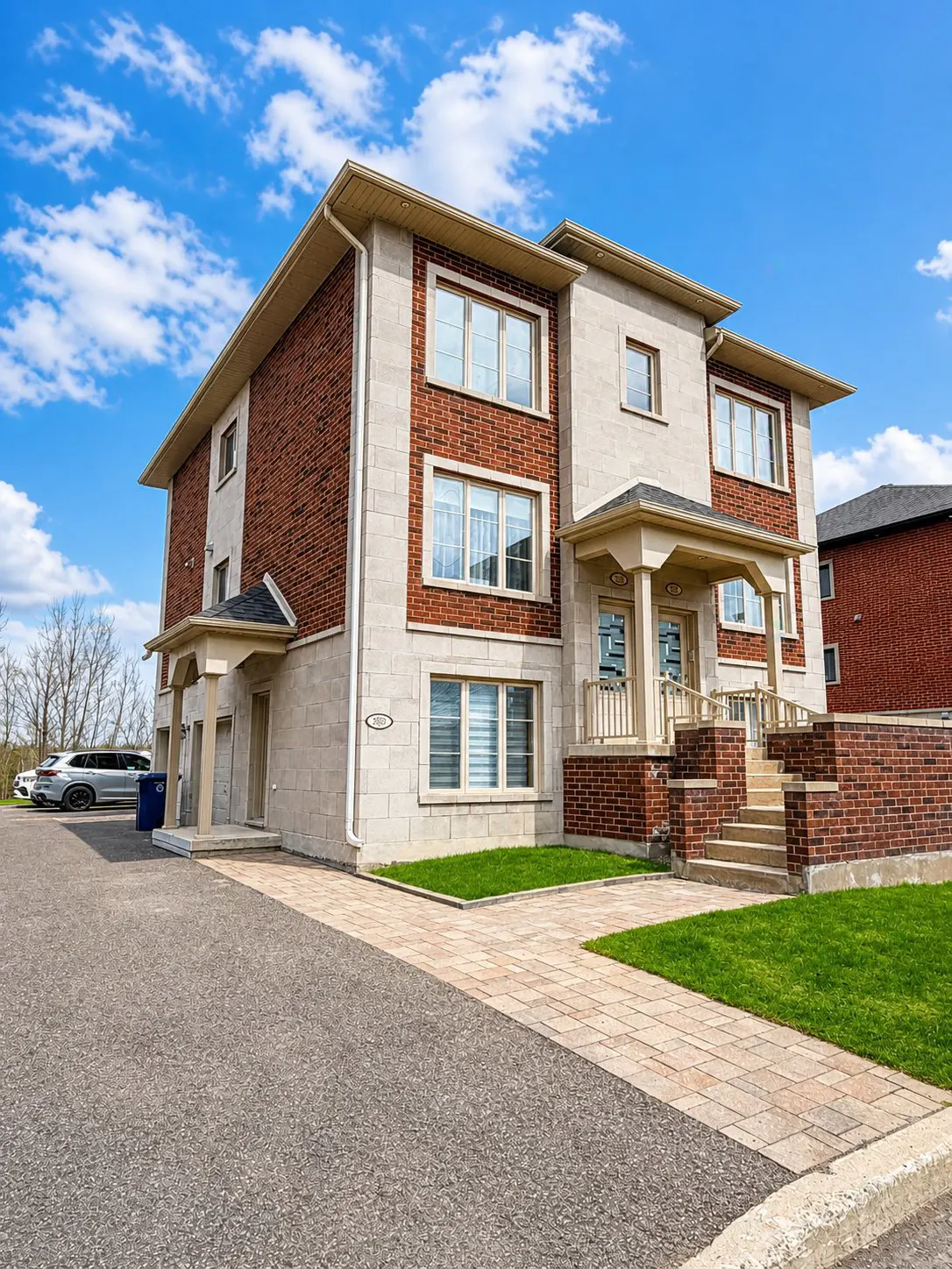 Two-story townhouse with brick and stone facade, front porch, and paved driveway under a blue sky.