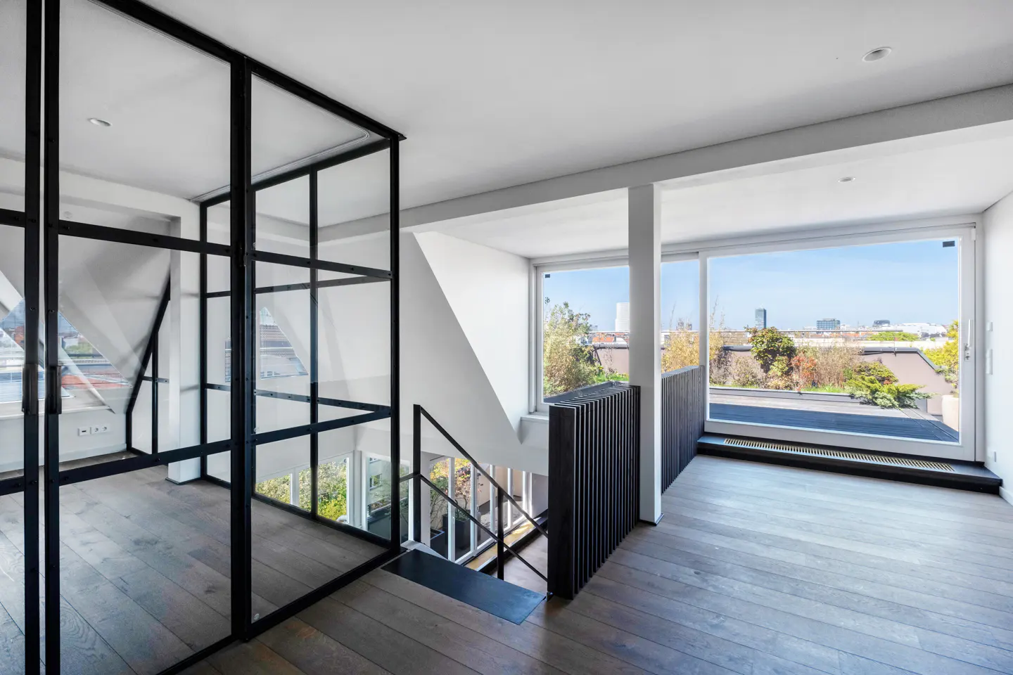 Bright, modern interior with wood floors, white walls, and black accents. A glass and metal partition separates the space, with a staircase visible. Large windows offer a city view.
