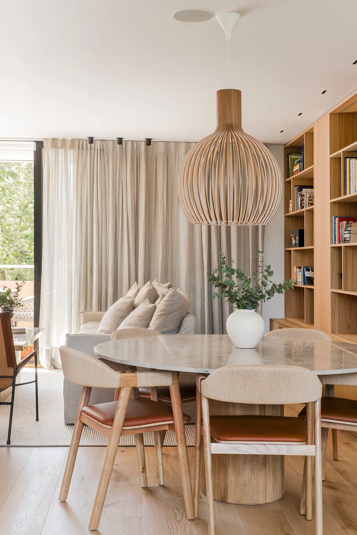 Bright living room with a round marble table, wooden chairs, and a unique wooden pendant light. Bookshelves and a sofa are in the background.