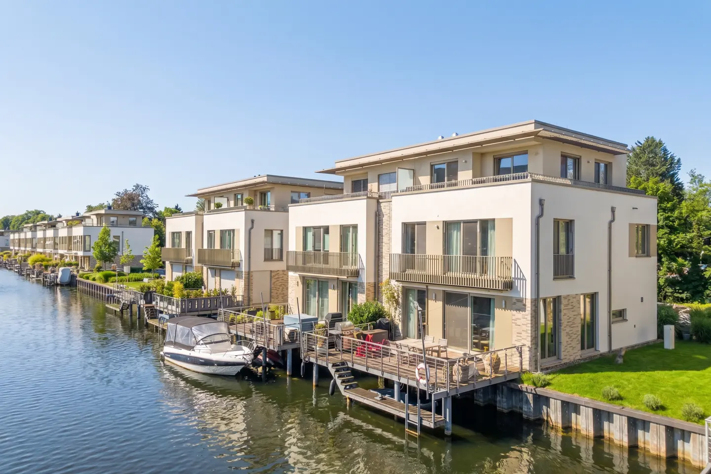Modern waterfront townhouses with docks on a canal under a clear blue sky. A white boat is docked in front.