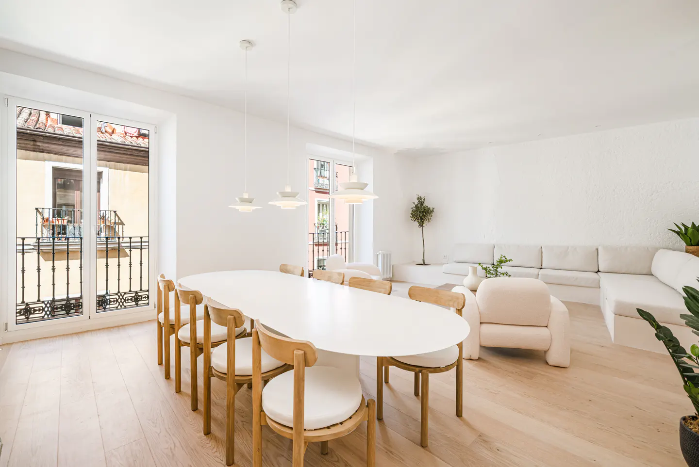 Bright, modern living room with white walls, light wood floors, and a large oval dining table with wooden chairs. A white sofa and plants add to the airy feel.