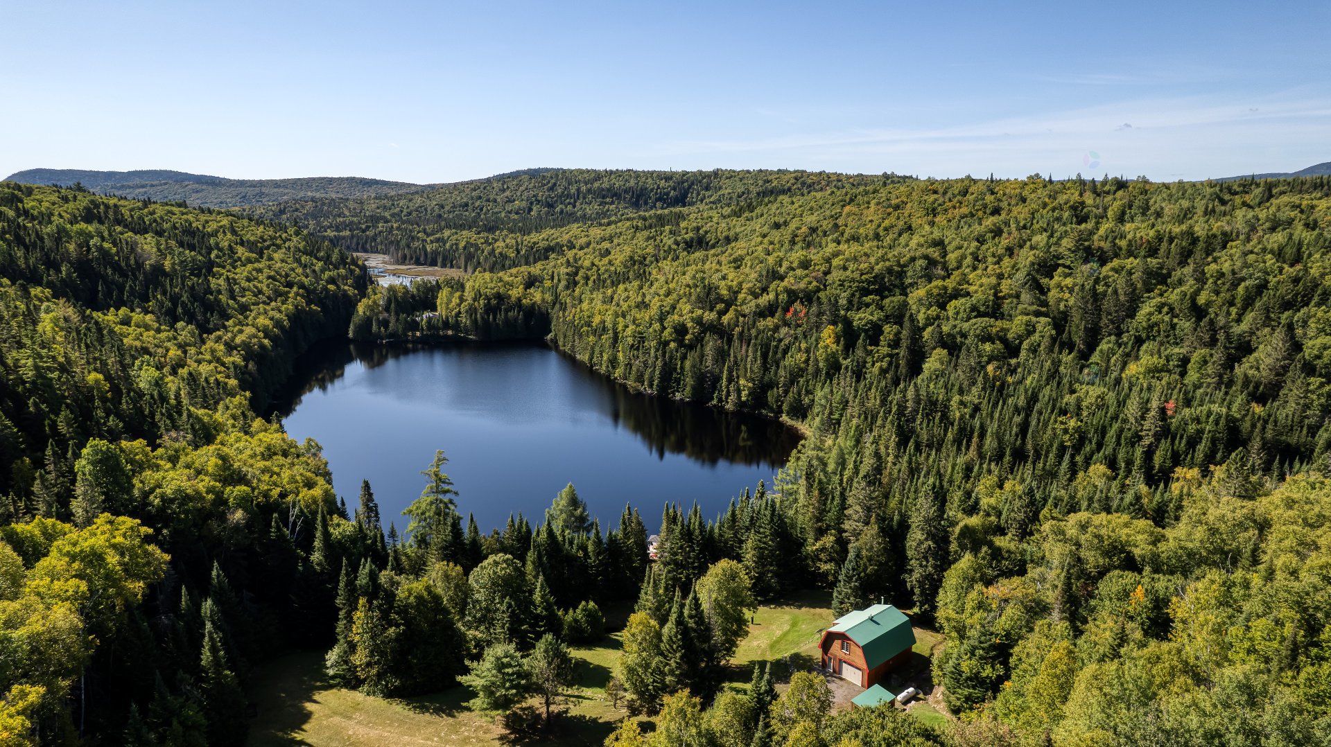 Aerial view of a dark blue lake surrounded by green forest and mountains under a clear blue sky. A small house with a green roof is visible near the lake.