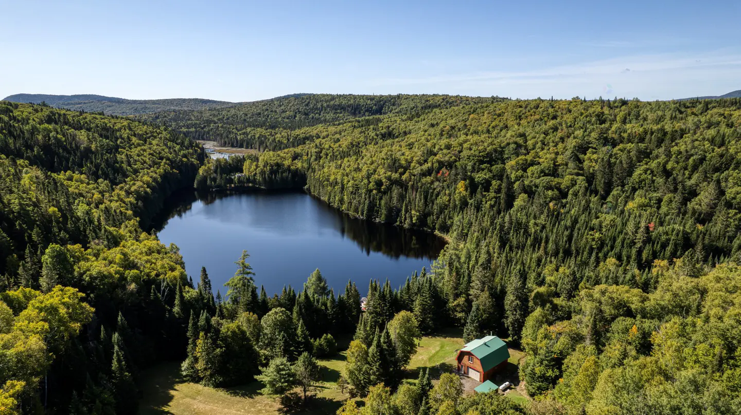 Aerial view of a dark blue lake surrounded by green forest and mountains under a clear blue sky. A small house with a green roof is visible near the lake.