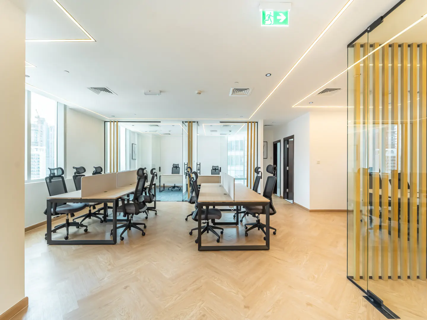 Bright office space with desks, black chairs, and wood floors. Glass partitions divide the room. An exit sign is visible on the ceiling.