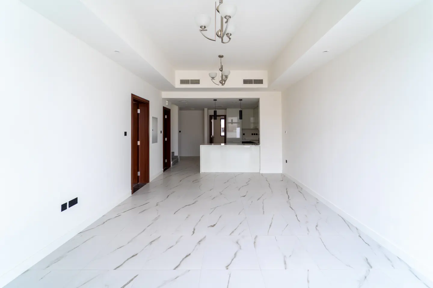 Bright, empty room with white walls, marble floors, and two chandeliers. A kitchen area is visible in the background.