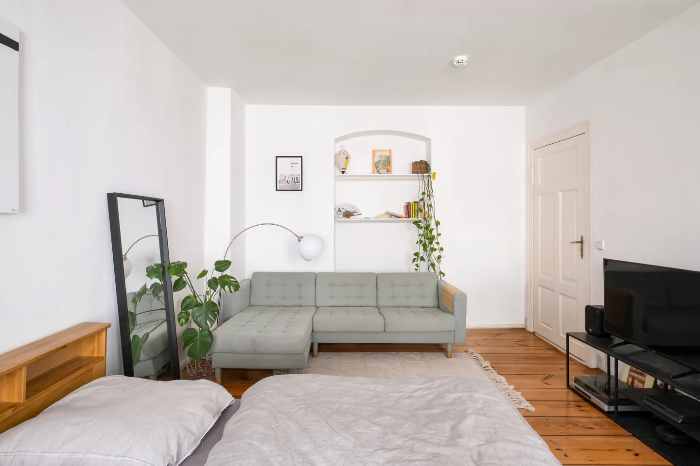Bright studio apartment with a gray sofa, bed, and wooden floors. A mirror and plant are near the bed. A built-in shelf is on the white wall.