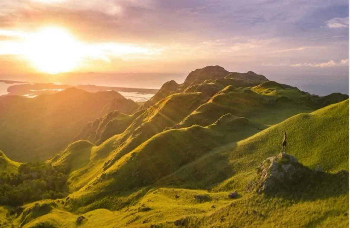 Scenic view of green rolling hills at sunset, with a person standing on a rock formation in the foreground.