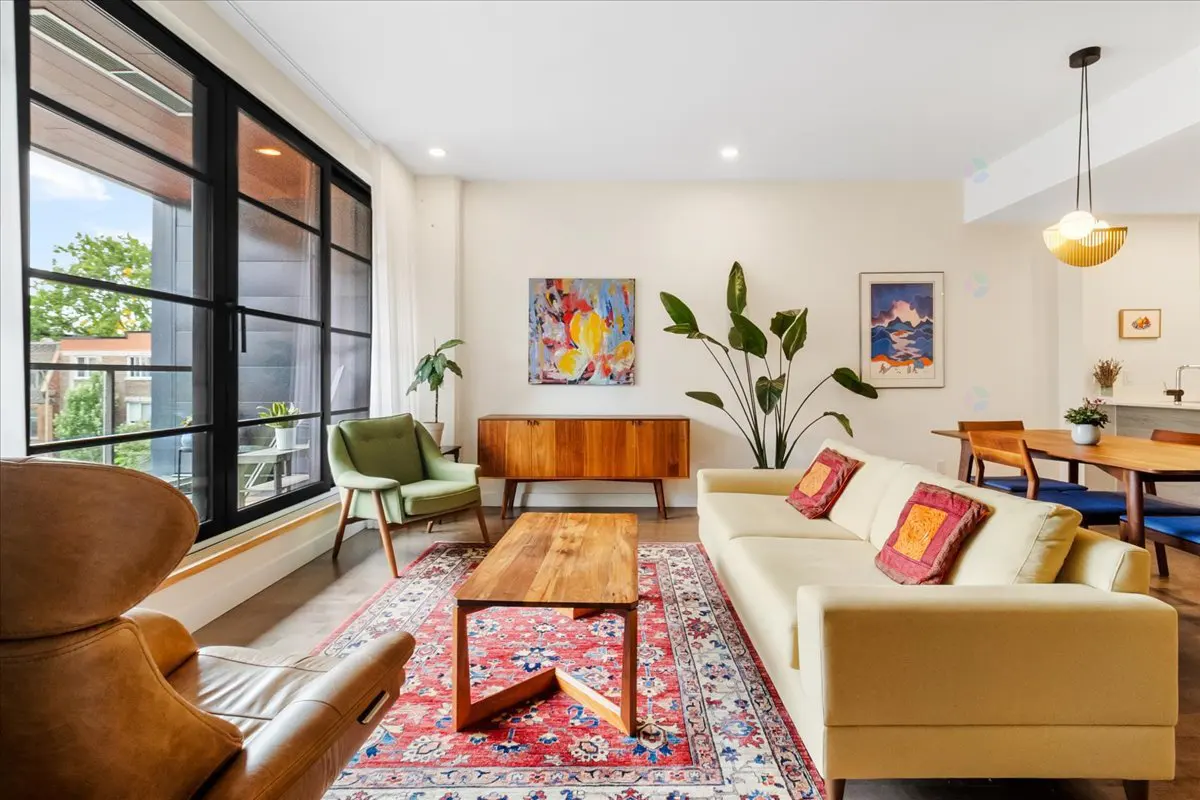 Bright living room with a tan sofa, wooden coffee table, and red patterned rug. Large black-framed windows let in natural light.