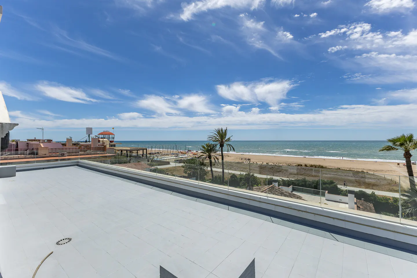 View from a rooftop overlooking a sandy beach, blue ocean, and palm trees under a partly cloudy sky.