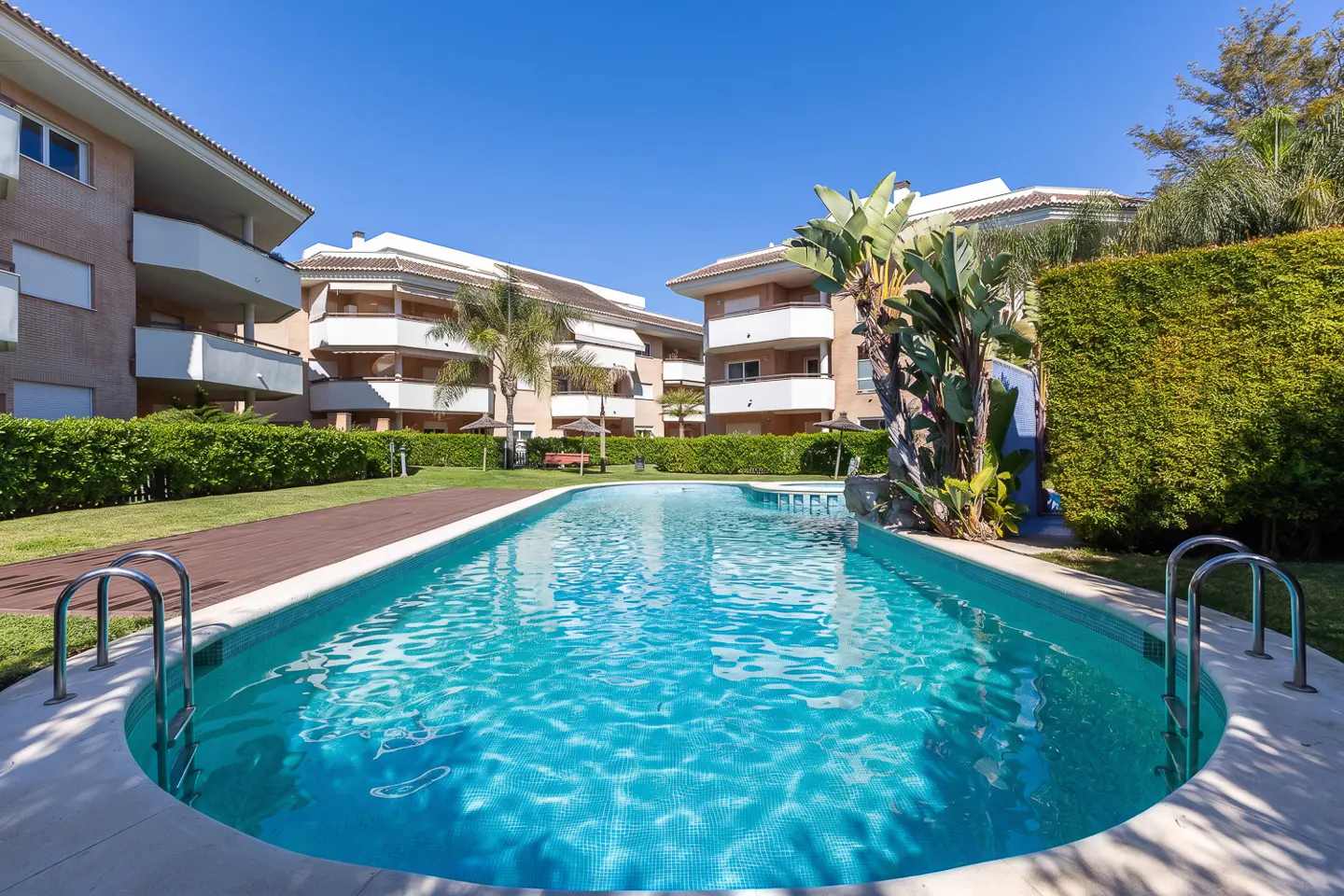 Outdoor pool with turquoise water, surrounded by apartments, green hedges, and a clear blue sky.