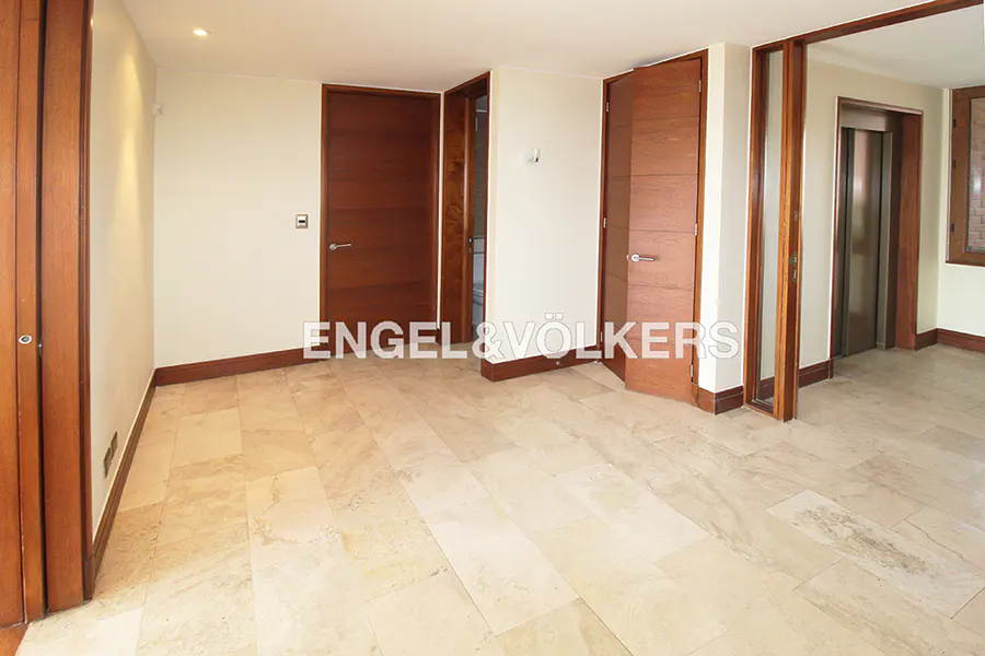An empty room with beige marble floors, cream walls, and dark wood trim. There are three doors and an elevator visible.