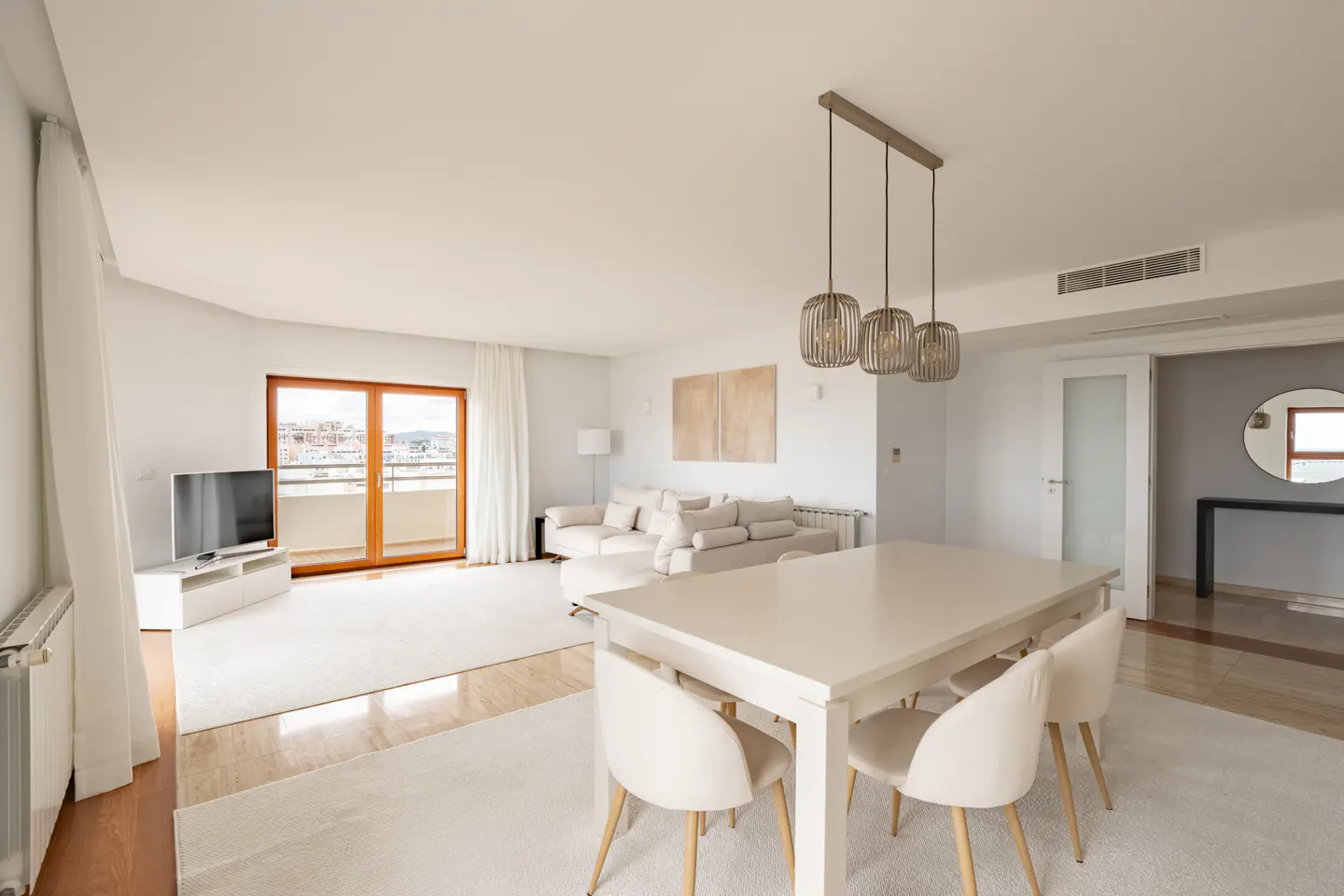 Bright, modern living and dining area with white walls, furniture, and a wood-framed balcony door. Three pendant lights hang above the dining table.